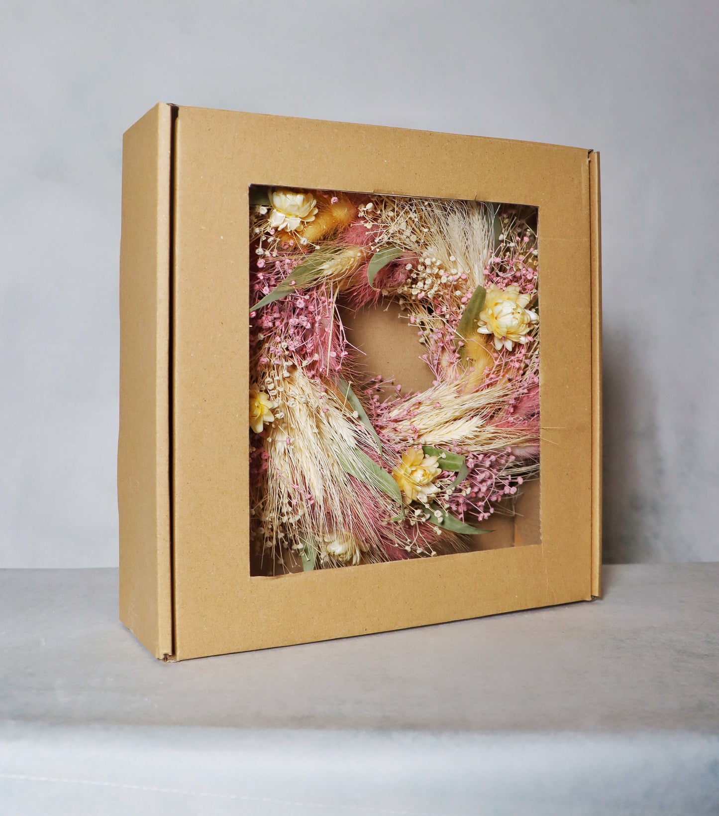 A cardboard box with a windowed front, displaying a circular dried flower wreath inside. The wreath features a mix of pink, yellow, and white dried flowers, along with green foliage and dried grasses, set on a grey background.