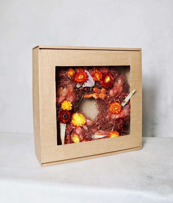 A cardboard box with a windowed front, displaying a circular dried flower wreath inside. The wreath features a vibrant mix of red, orange, and yellow dried flowers, along with purple and green foliage accents on a plain background.