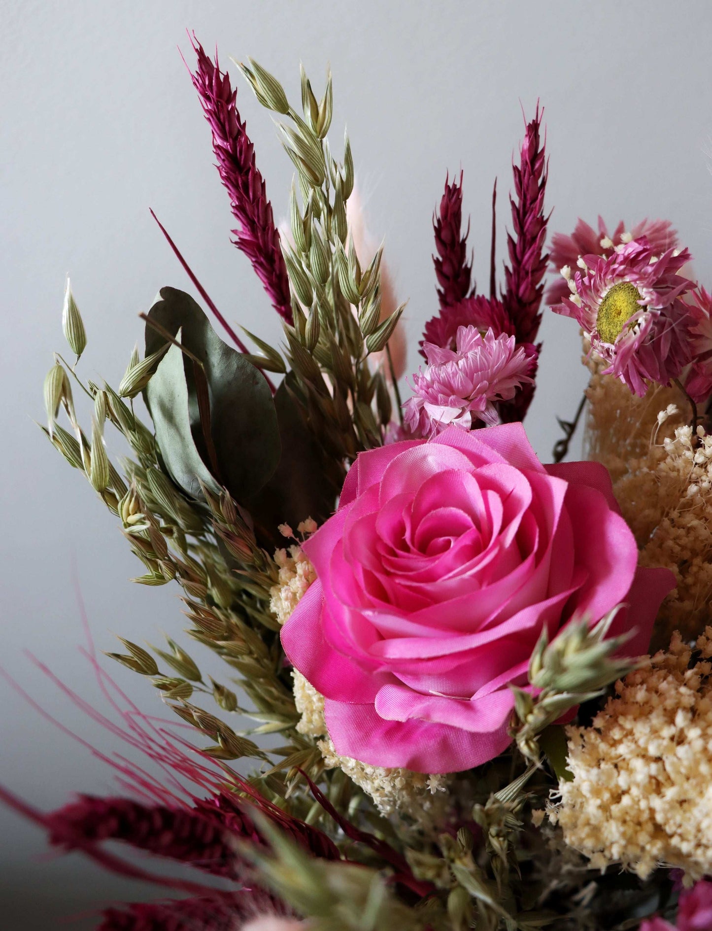 A close-up image of a dried and artificial flower bouquet with a pink rose, wheat and other beige and pink flowers.