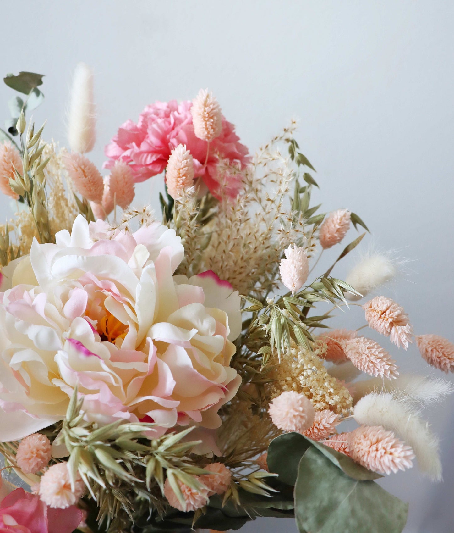 A close-up of a floral arrangement featuring large pink and white peonies, smaller pink blooms, and various dried grasses and foliage, displayed against a light grey background.