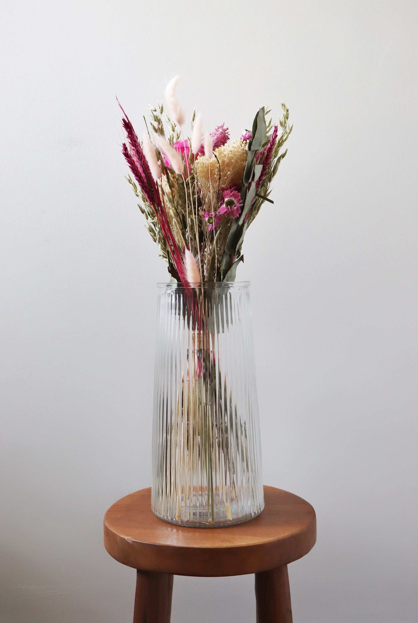 Dried flower bouquet with pink, beige and purple flowers in a ribbed glass vase on a wooden stool, with a white background.
