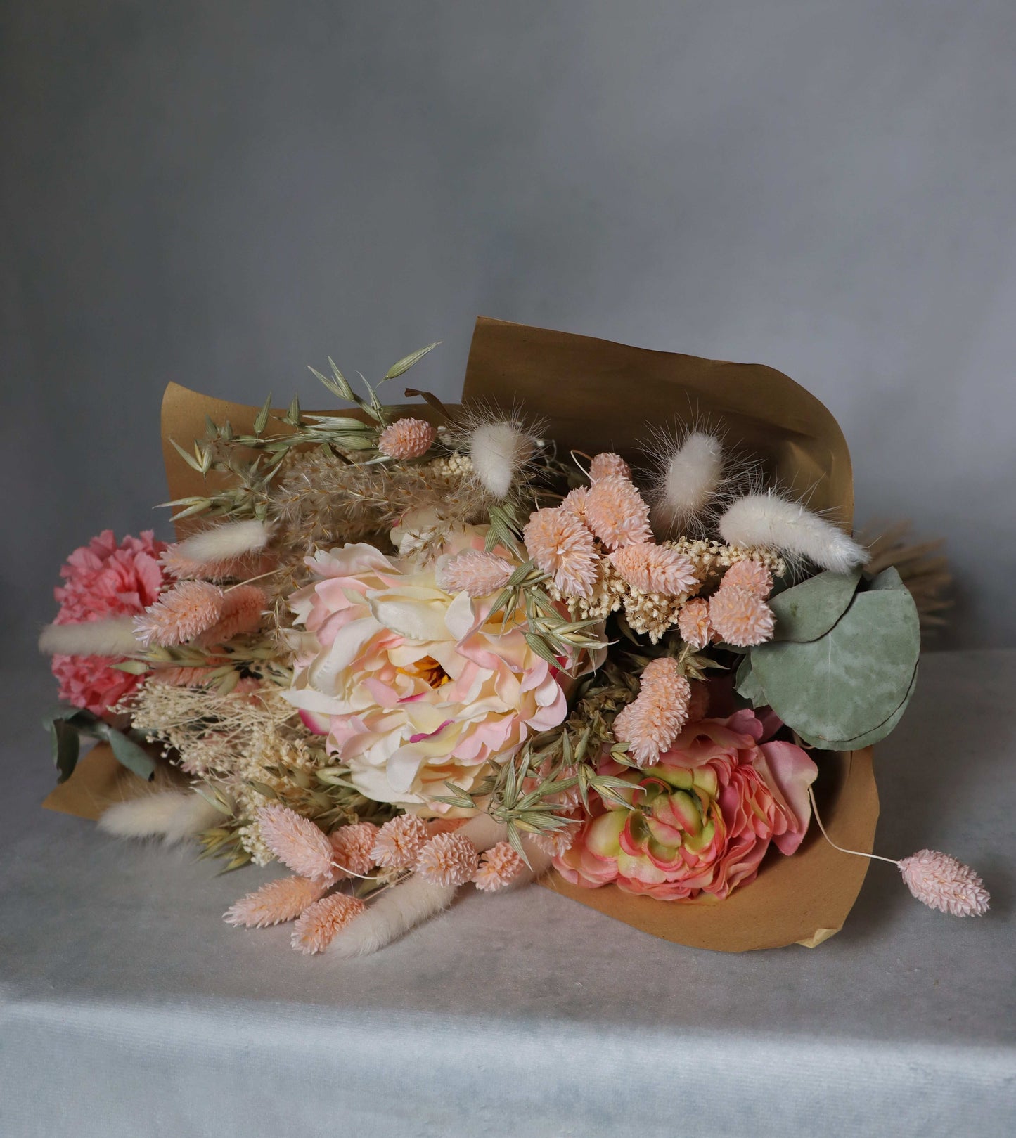 A bouquet of dried flowers wrapped in brown paper, featuring pink, peach, and white blooms, along with dried grasses and green foliage. The bouquet is placed on a surface against a grey background.