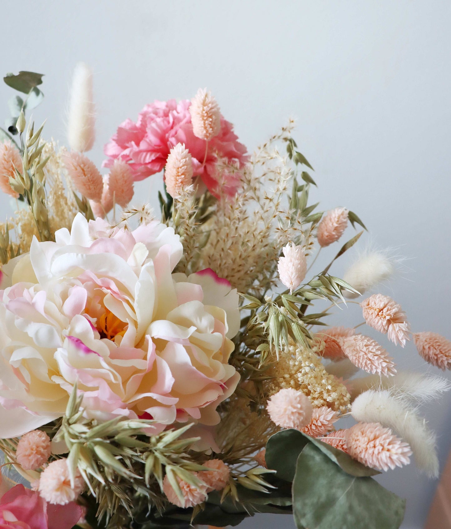 A close-up of a floral arrangement featuring large pink and white peonies, smaller pink blooms, and various dried grasses and foliage in shades of green and beige, displayed against a light grey background.