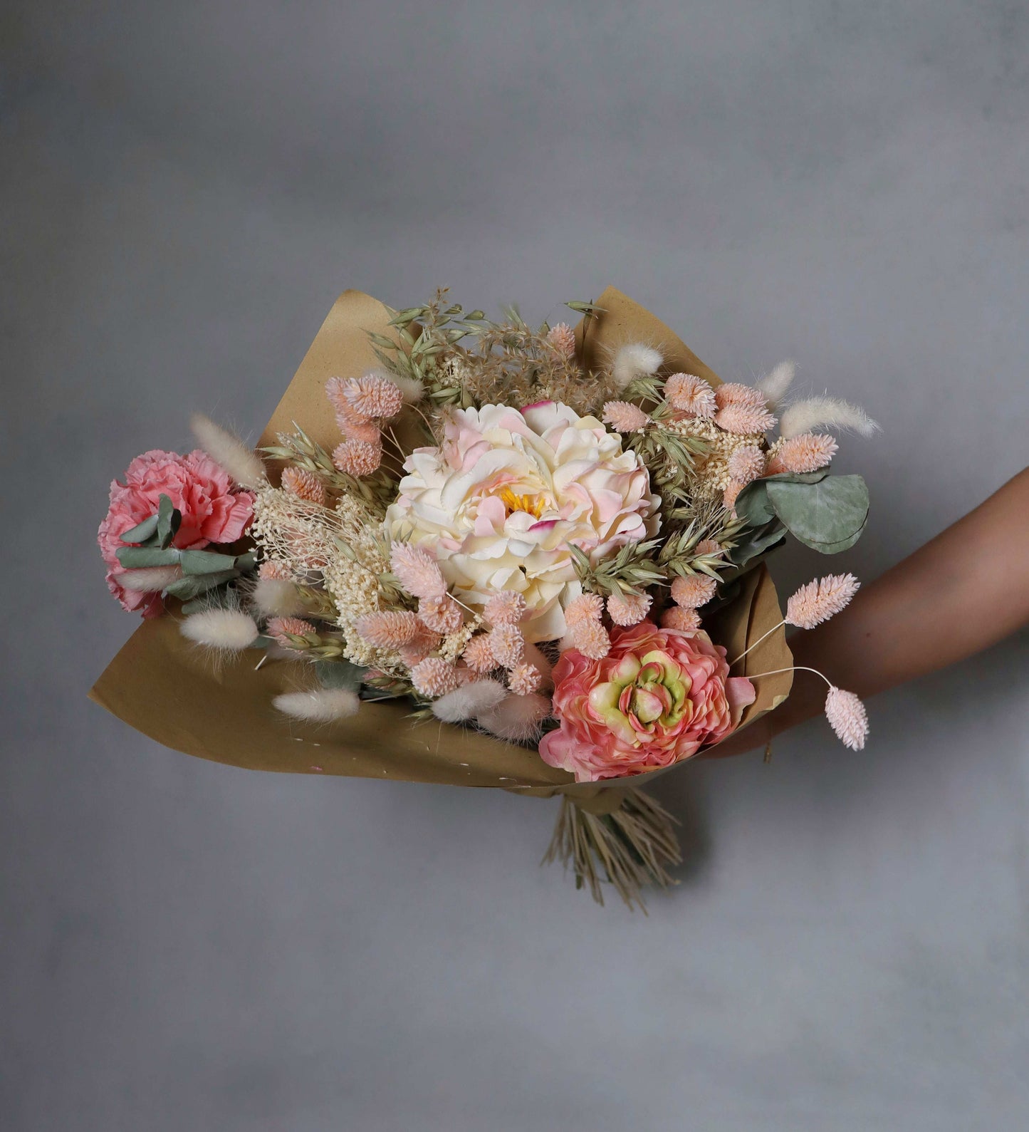 A bouquet of dried flowers wrapped in brown paper, featuring pink, peach, and white blooms, along with dried grasses and green foliage. The bouquet is held in a person's hand against a grey background.