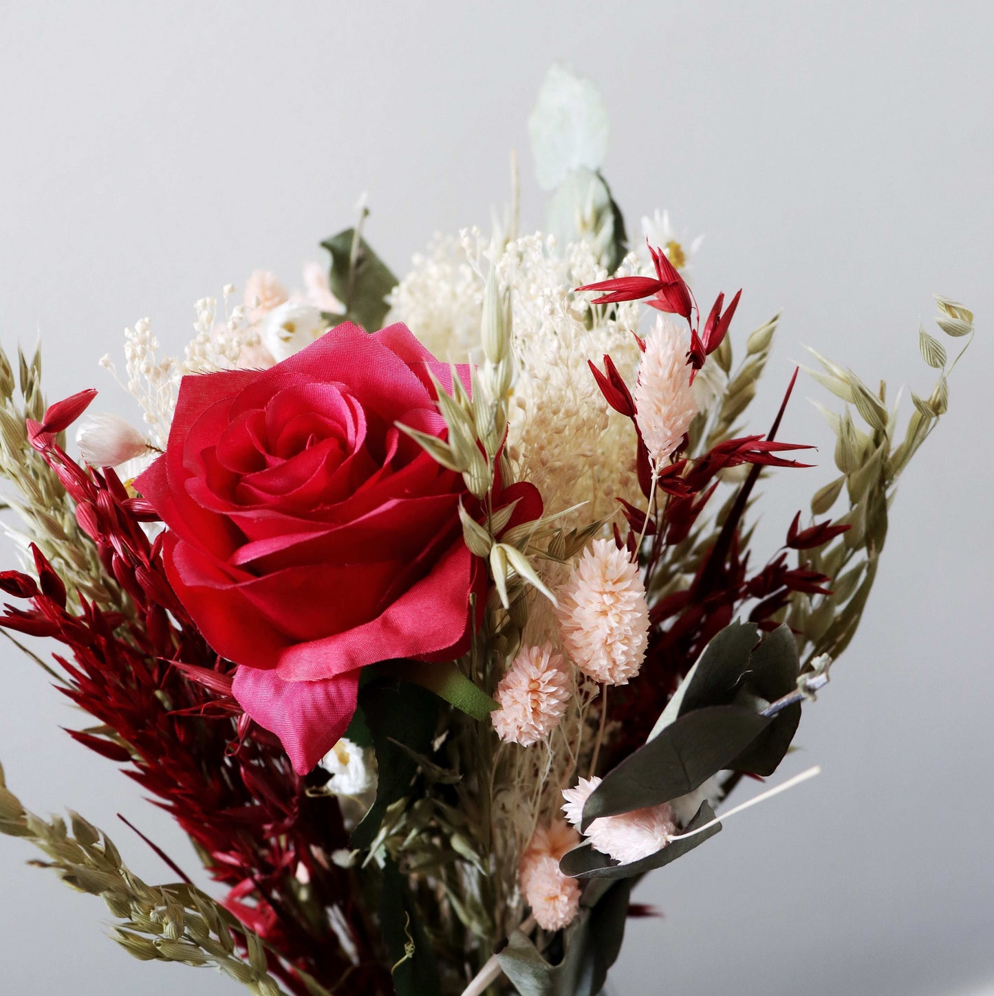 Close up of dried flowers with a red rose, pink blooms, white baby's breath, and green eucalyptus leaves against a grey background.