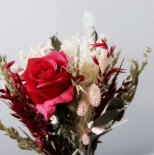 Close up of dried flowers with a red rose, pink blooms, white baby's breath, and green eucalyptus leaves against a grey background.