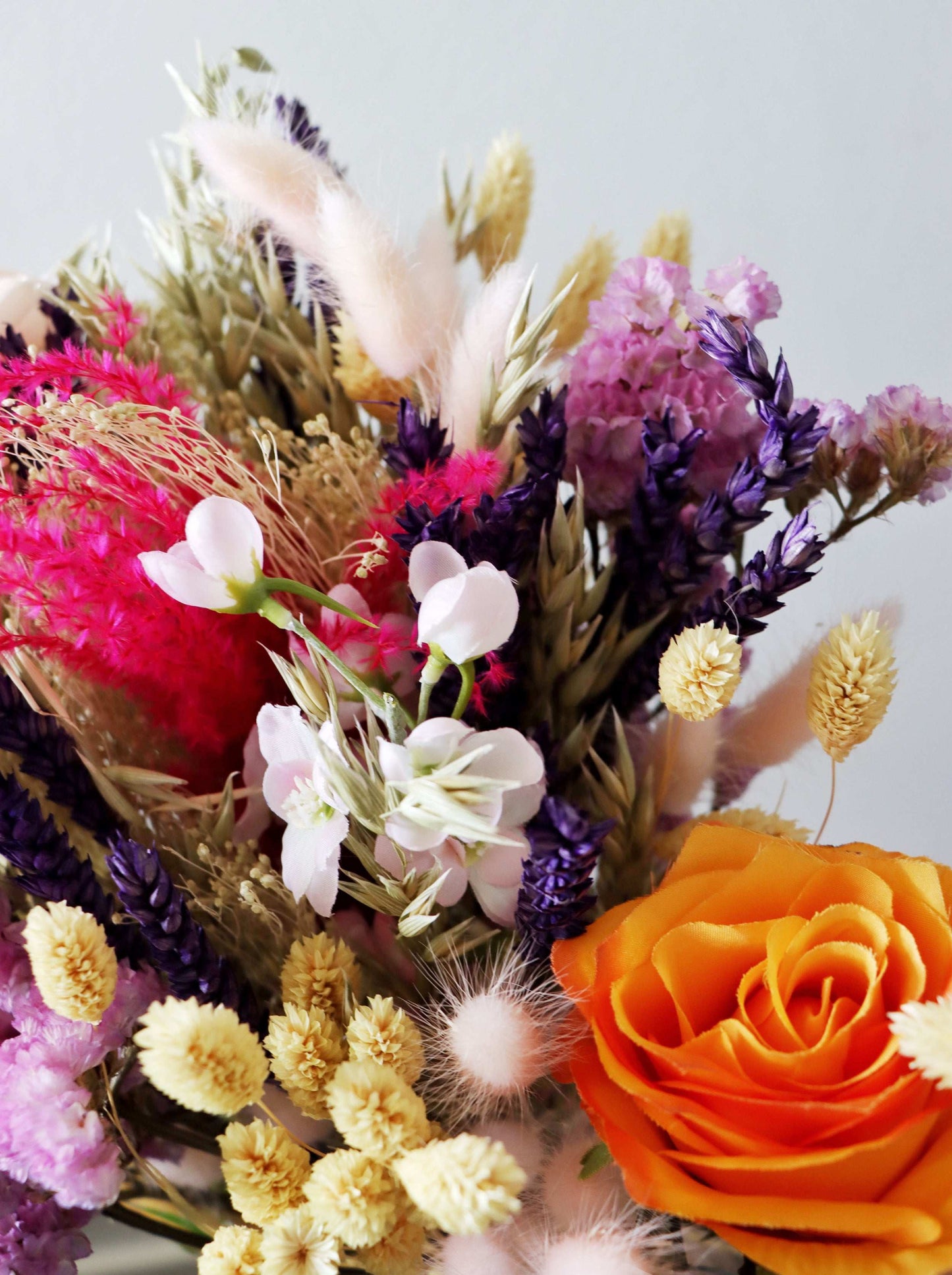 A close-up of a vibrant dried flower arrangement featuring pink, purple, red, and orange blooms, along with beige and yellow stems and fluffy accents, set against a white background.