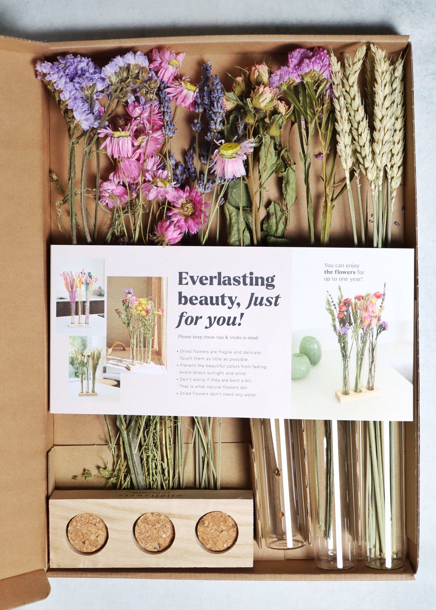 Open box with dried flowers in purple, pink, and yellow, glass tubes, and a wooden stand with corks on a white background.