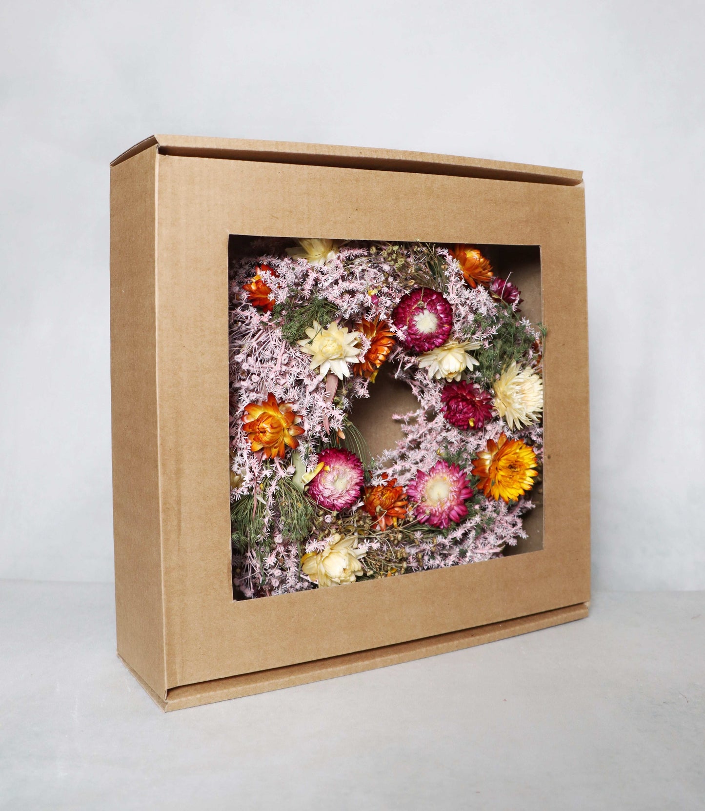 A cardboard box with a windowed front, showcasing a circular dried flower wreath inside. The wreath features a mix of pink, orange, yellow, red, and white dried flowers, along with green foliage, displayed against a white background.