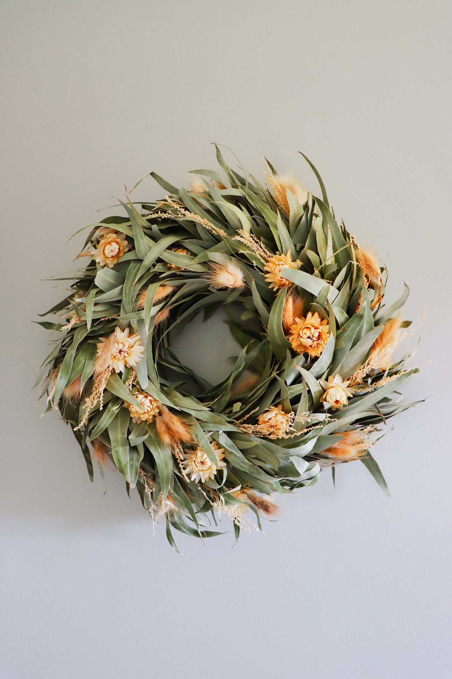 A circular wreath made of dried flowers and foliage, featuring light orange blooms and green leaves, against a light grey background.