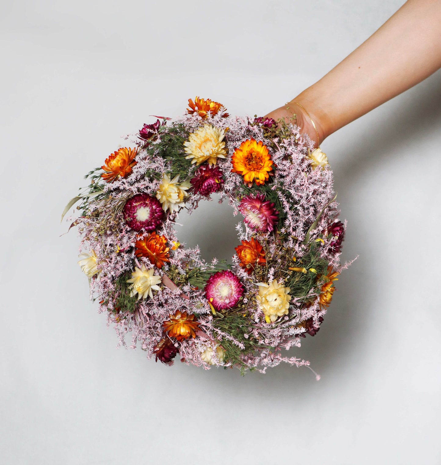 A circular wreath made of dried flowers, featuring a vibrant mix of pink, orange, yellow, red, and white blooms, along with delicate green foliage. The wreath is held in a person's hand on a white background.