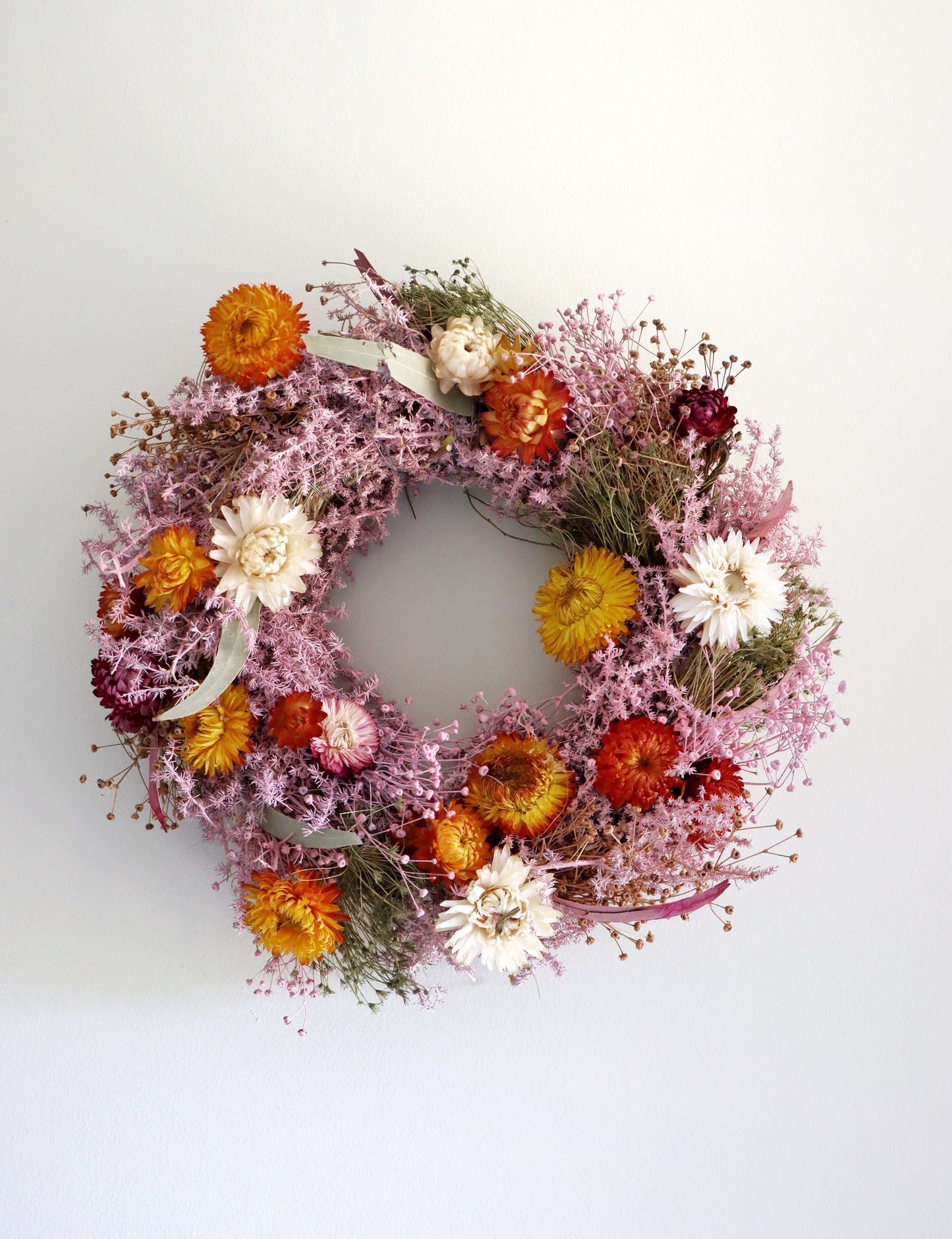 A circular wreath made of dried flowers, featuring a mix of pink, orange, red, and white blooms displayed against a plain white background.