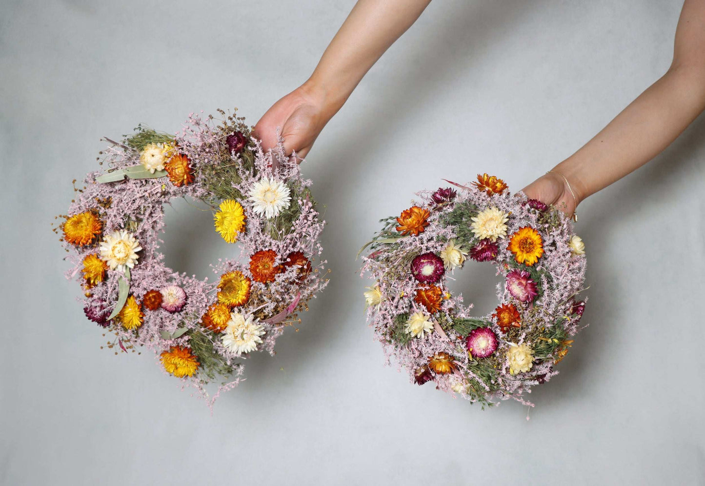 Hands holding a large and small circular wreath made of dried flowers with purple, yellow, and pink blooms and green foliage against a white background.