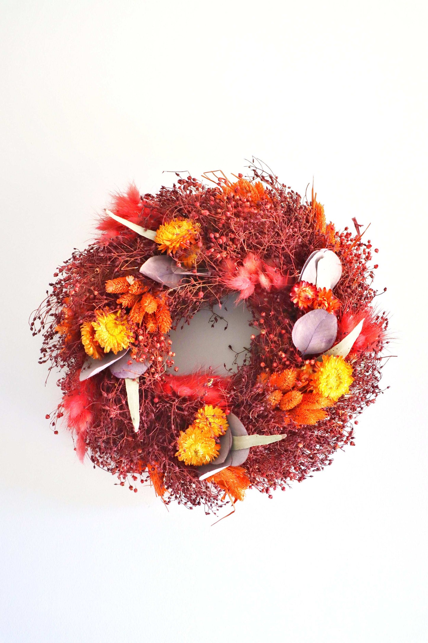 A circular wreath made of dried flowers, featuring a rich mix of red, orange, and yellow blooms, along with purple foliage accents and delicate dried grasses. The wreath is against a plain white background.