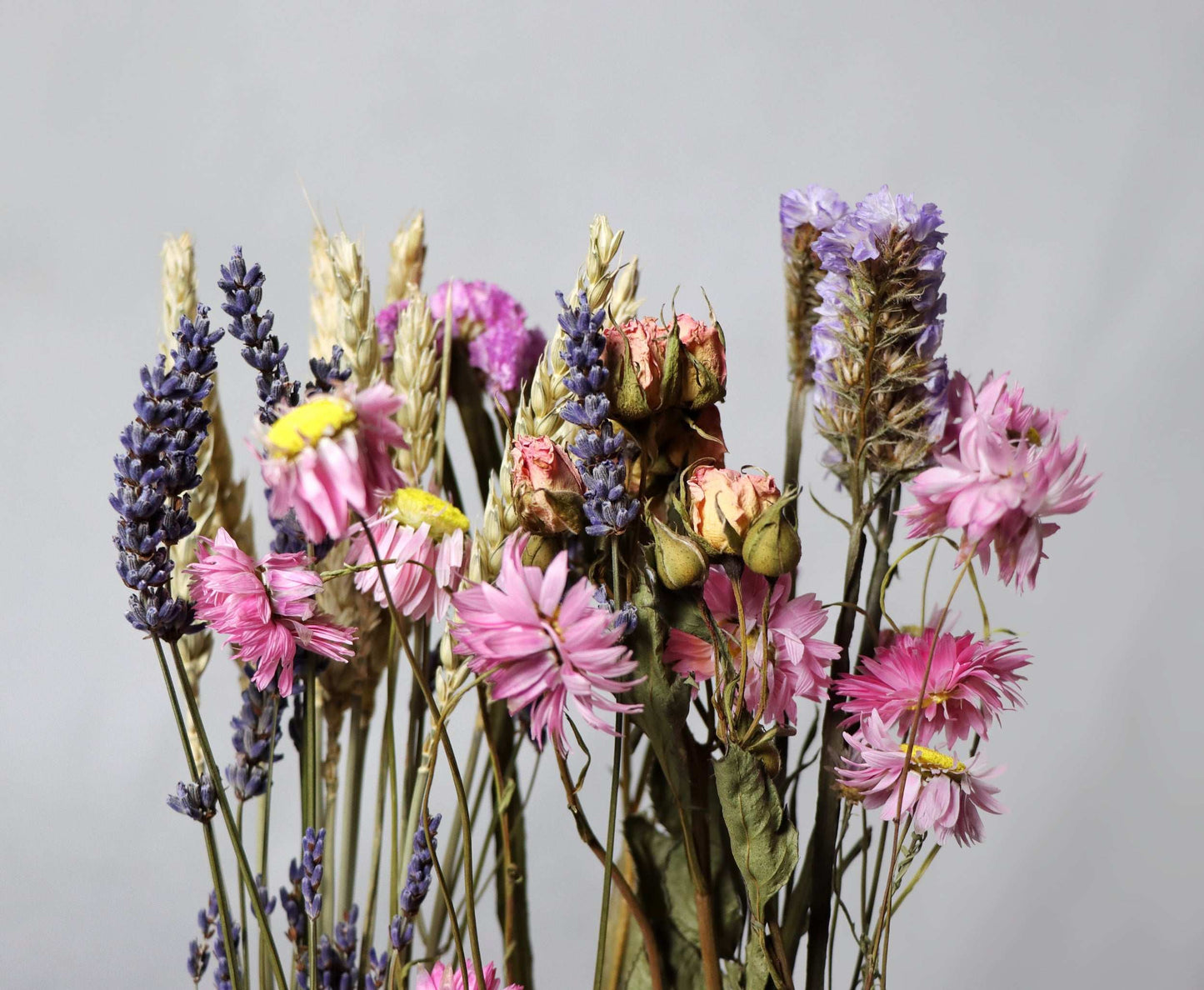 Bunch of dried flowers with pink roses, purple lavender, yellow daisies, and wheat stalks against a white background.