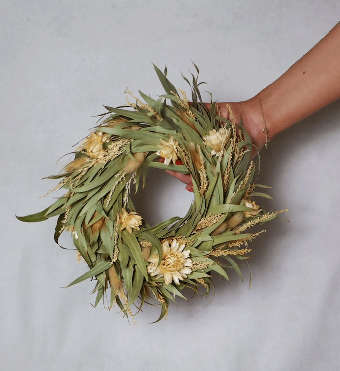 A circular wreath made of dried flowers and foliage, featuring light yellow blooms and green leaves. The wreath is held in a person's hand against a plain grey background.