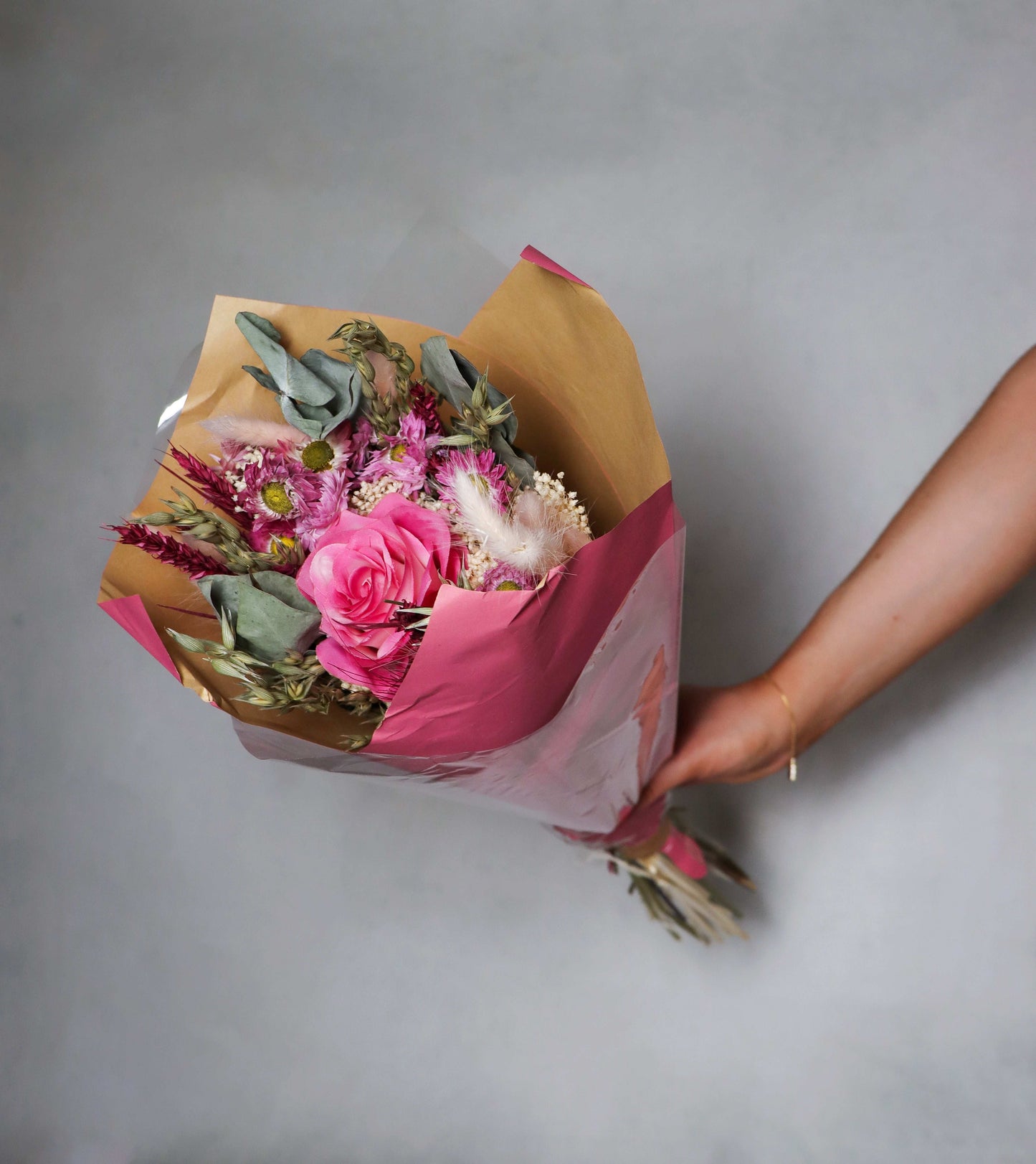 Bouquet of dried flowers wrapped in brown and pink paper held by a hand on a grey background.