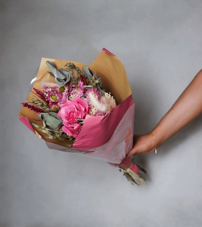 Bouquet of dried flowers wrapped in brown and pink paper held by a hand on a grey background.