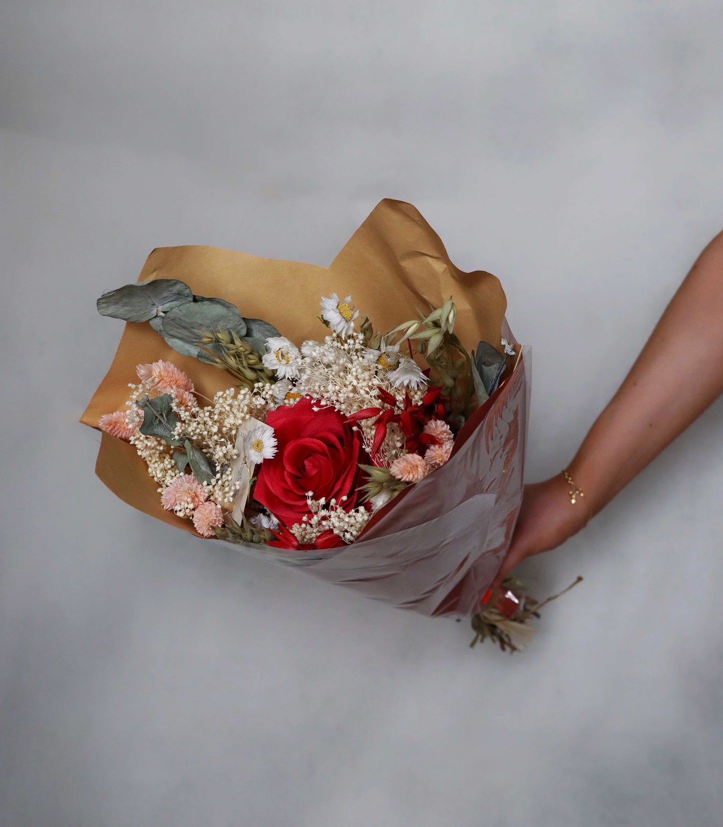 Hand holding a bouquet of dried flowers with pink flowers and red roses, eucalyptus, and baby’s breath wrapped in brown paper against a grey background.