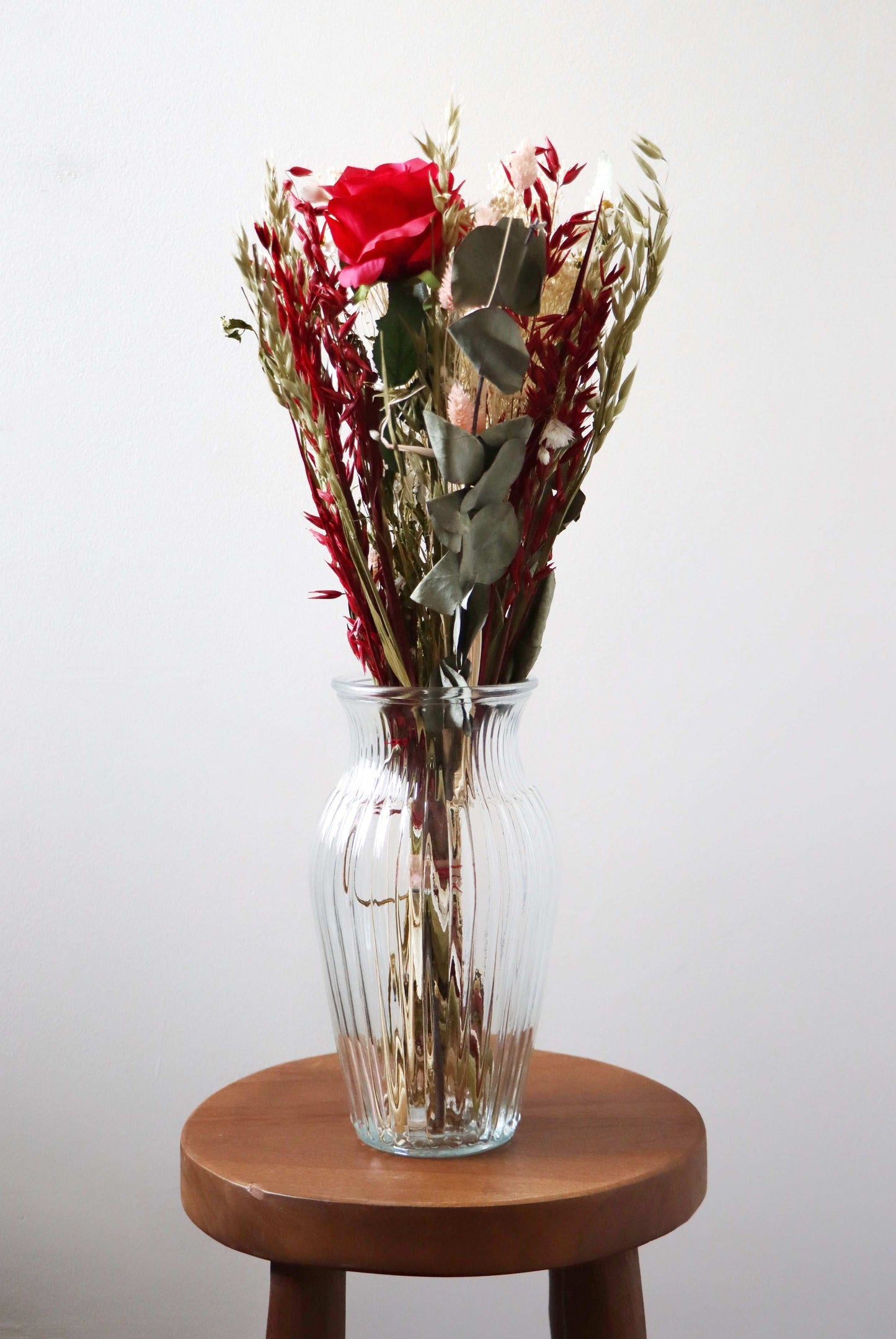 Clear ribbed glass vase with dried red roses, wheat stalks, and green leaves on a wooden stool against a white background.