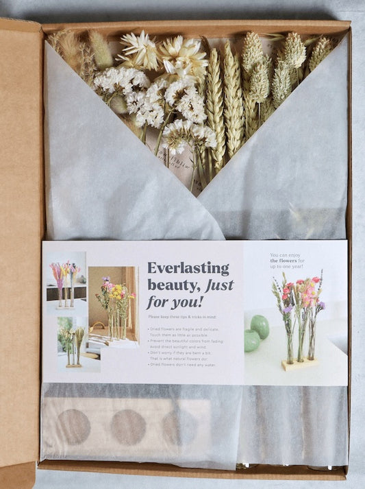 Three glass tubes with dried white and beige flowers, pampas grass, and wheat stalks on a wooden stand against a grey background.