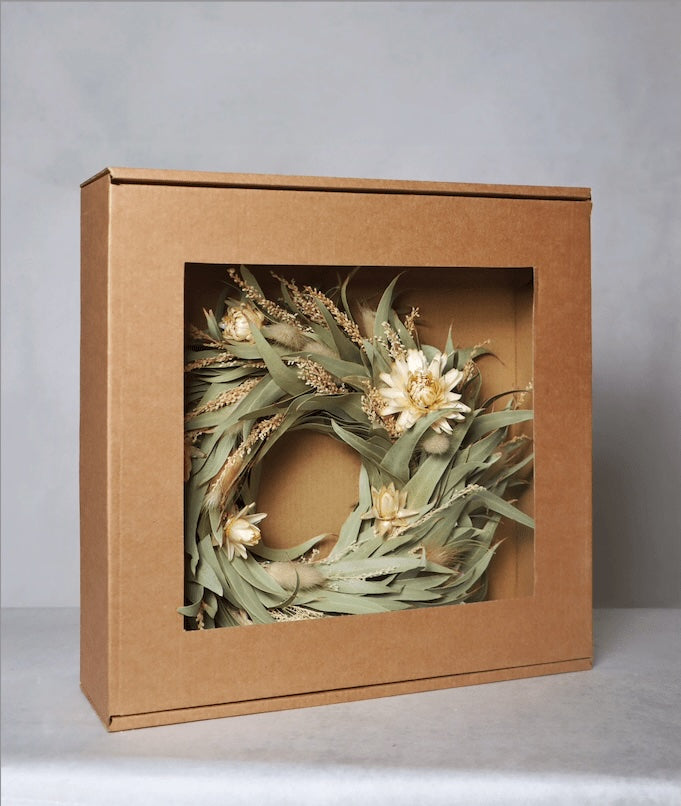 A cardboard box with a windowed front, displaying a circular dried flower wreath inside. The wreath features light yellow blooms and green foliage, set against a grey background.