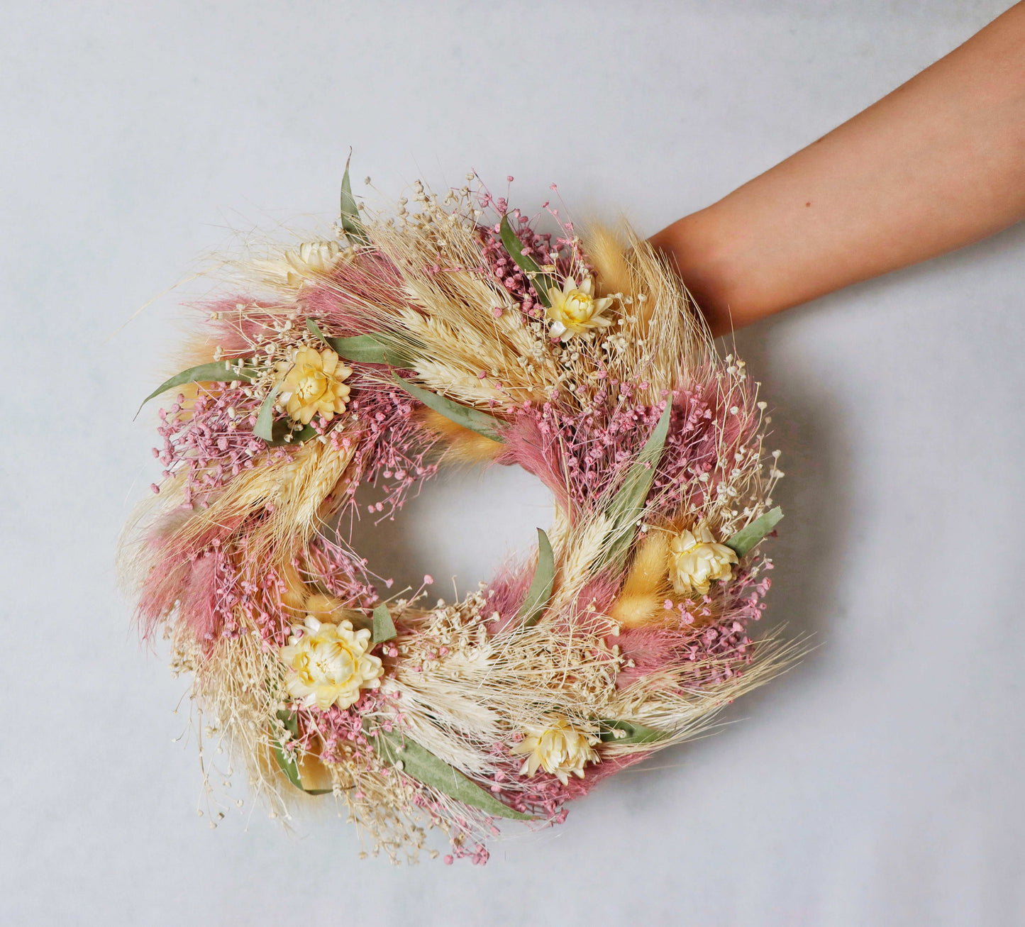 A circular wreath made of dried flowers and foliage, featuring pink, yellow, and white blooms, along with tan grasses and green leaves. The wreath is hand held against a grey background.