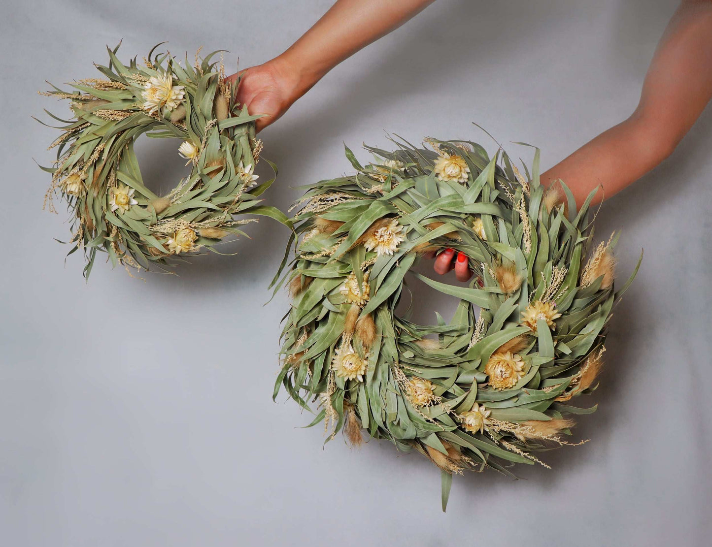 Two circular wreaths made of dried flowers and foliage, featuring light yellow blooms and green leaves. The wreaths are held in a person's hands against a plain grey background.