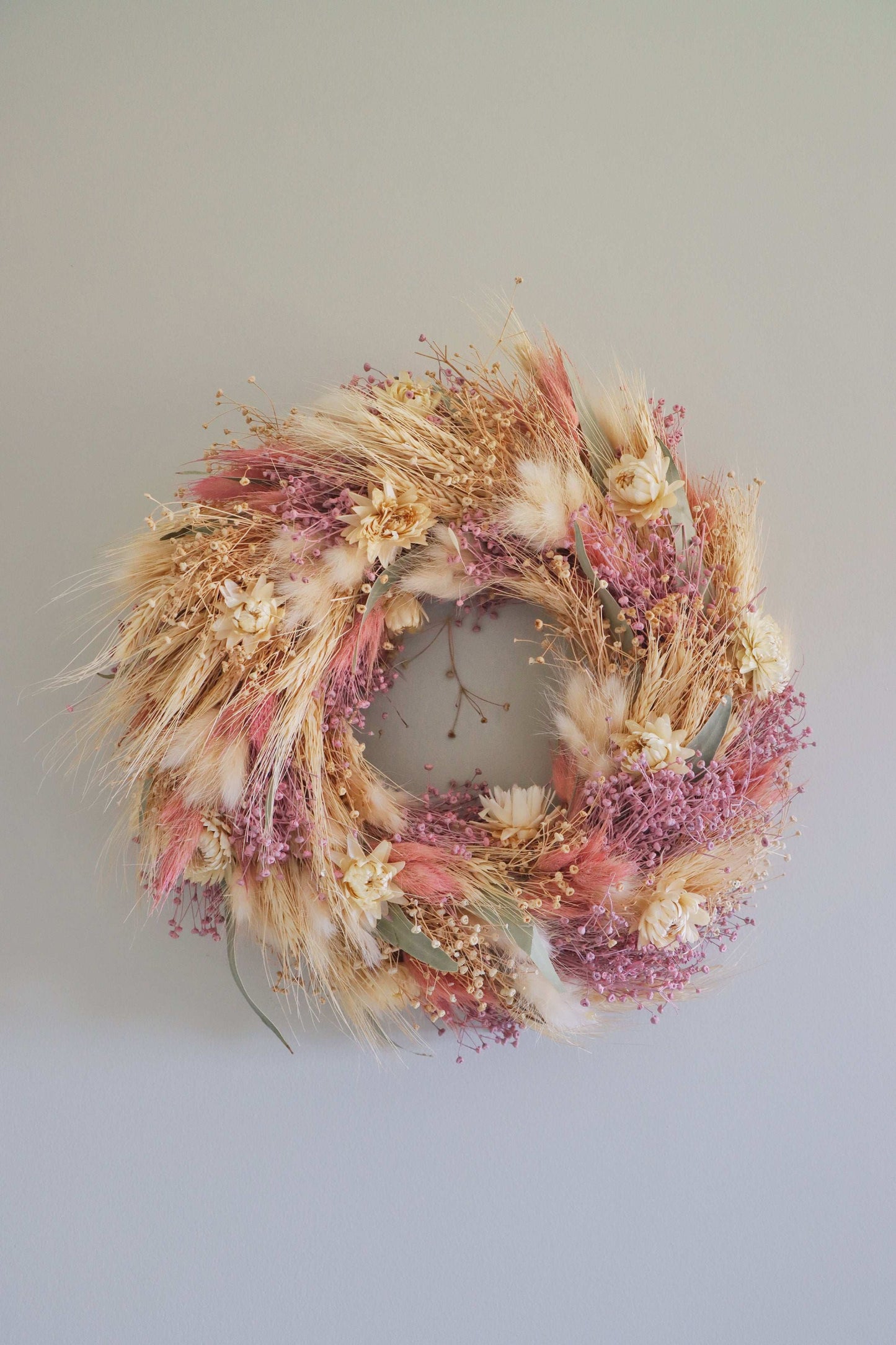 A circular wreath made of dried flowers and foliage, featuring pink, beige, and white blooms, along with green and tan grasses. The wreath is displayed against a white background.