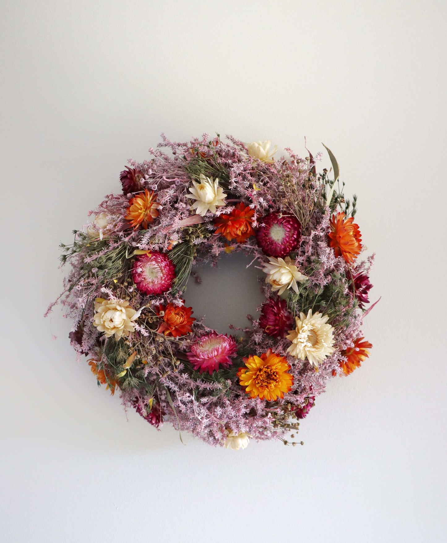 A circular wreath made of dried flowers, featuring a vibrant mix of pink, orange, red, and white flowers. The wreath is displayed against a plain white background.
