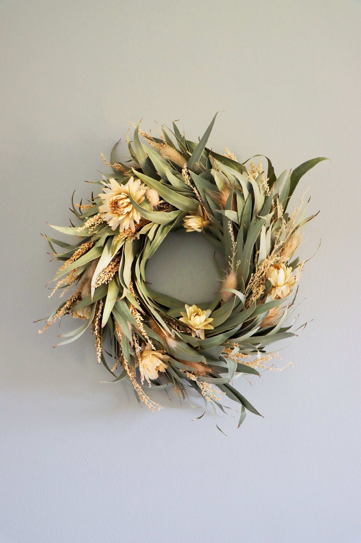 A circular wreath made of dried flowers and foliage, featuring light yellow blooms and green leaves, displayed against a light grey background.