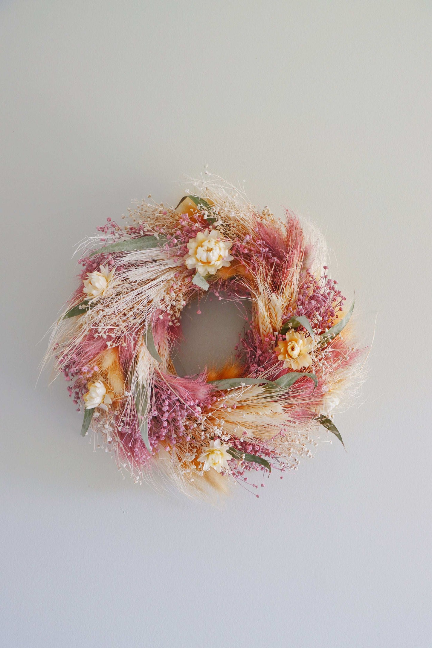 A circular wreath made of dried flowers and foliage, featuring pink, yellow, and white blooms, along with green leaves and tan grasses. The wreath is displayed against a white background.