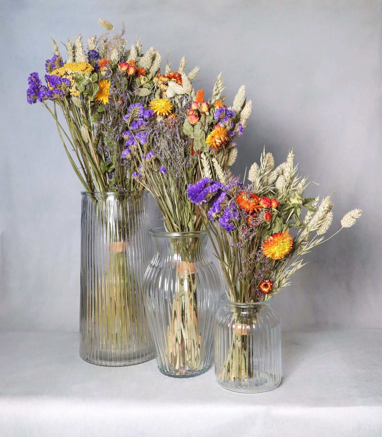 Three glass vases of varying sizes, each filled with dried flowers including purple statice, yellow and orange strawflowers, and dried grasses against a grey background.