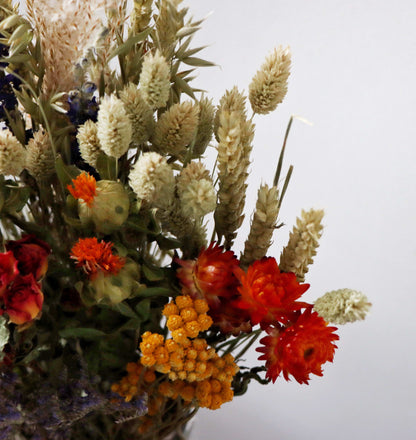 A bouquet of dried flowers in a vase, featuring red and orange strawflowers, yellow billy buttons, and dried grasses against a plain white background.