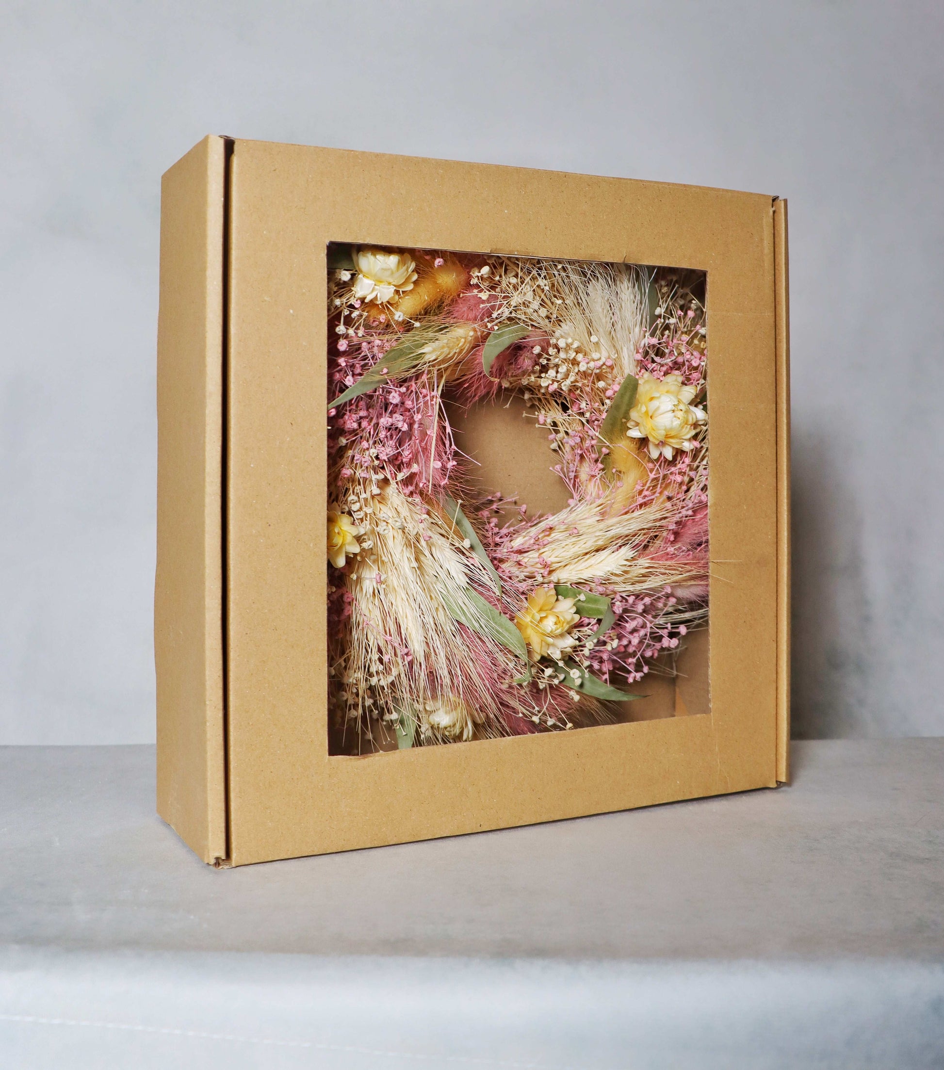 A cardboard box with a windowed front, displaying a circular dried flower wreath inside. The wreath features a mix of pink, yellow, and white dried flowers, along with green foliage and dried grasses, set on a grey background.