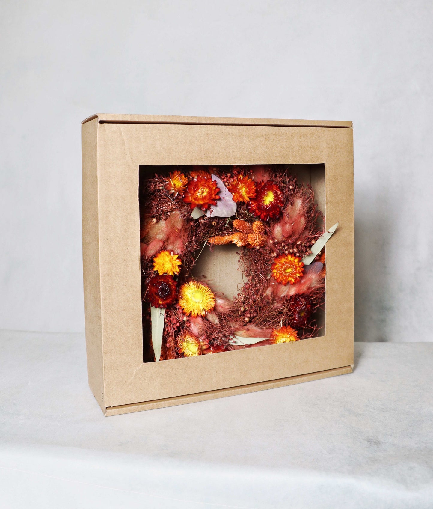 A cardboard box with a windowed front, displaying a circular dried flower wreath inside. The wreath features a vibrant mix of red, orange, and yellow dried flowers, along with purple and green foliage accents on a plain background.