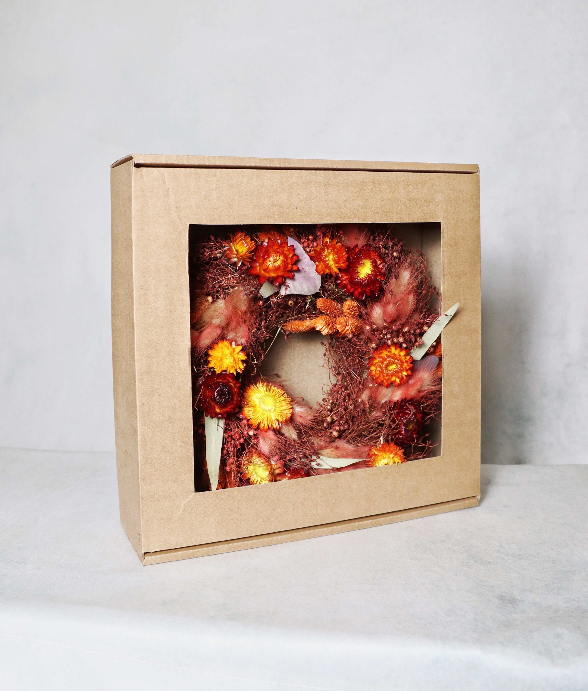 A cardboard box with a windowed front, displaying a circular dried flower wreath inside. The wreath features a vibrant mix of red, orange, and yellow dried flowers, along with purple and green foliage accents on a plain background.