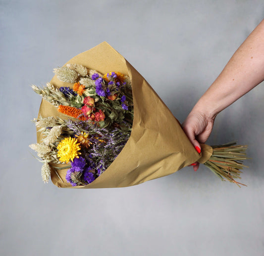 A colourful bouquet of dried flowers wrapped in brown paper, featuring purple statice, yellow strawflowers, orange celosia, and dried grasses, held by a hand against a grey background.
