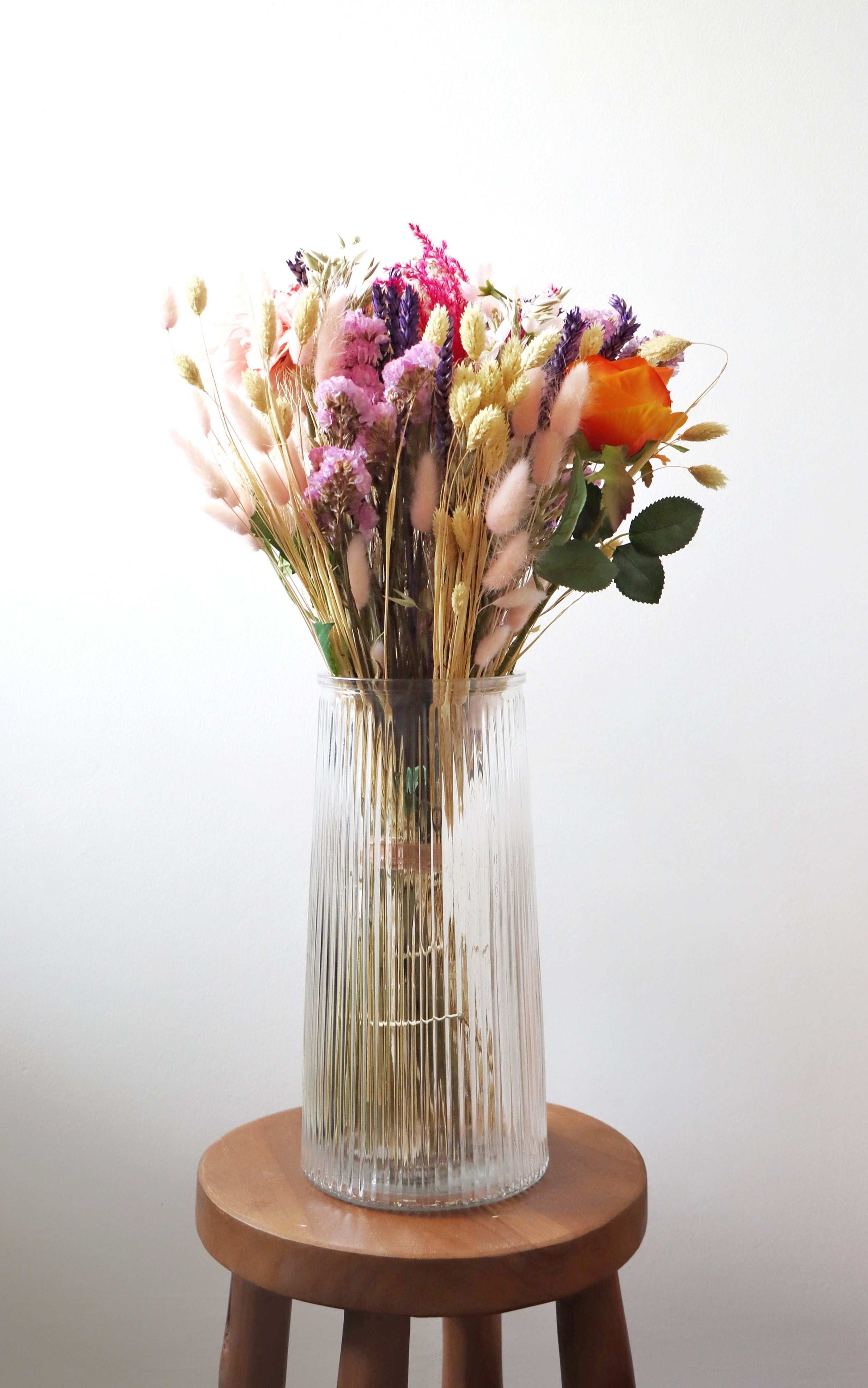 A tall, clear glass vase filled with dried flowers, including pink, purple, and orange blooms, along with beige and green stems and leaves, placed on a wooden stool against a white background.