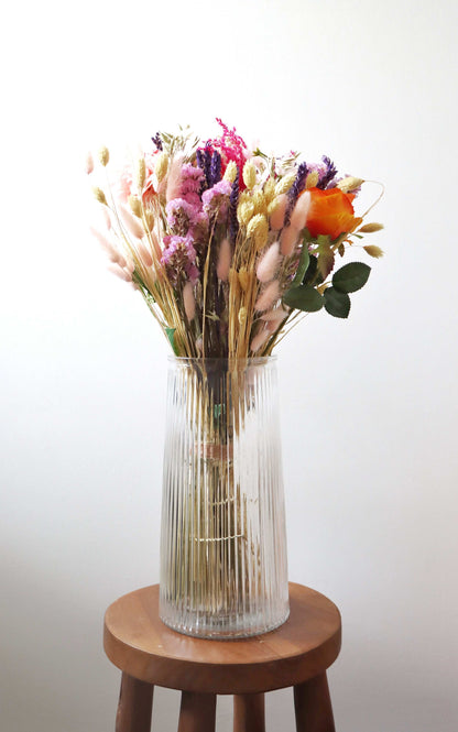 A tall, clear glass vase filled with dried flowers, including pink, purple, and orange blooms, along with beige and green stems and leaves, placed on a wooden stool against a white background.