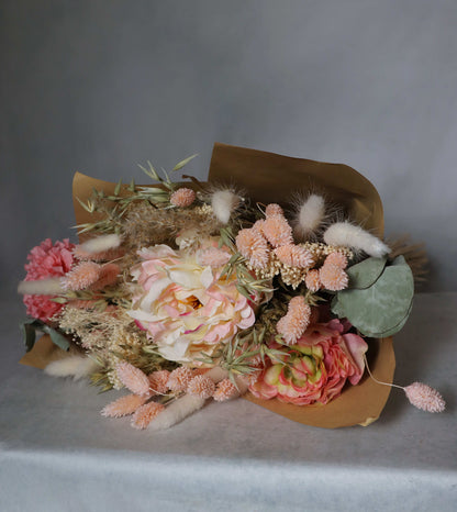 A bouquet of dried flowers wrapped in brown paper, featuring pink, peach, and white blooms, along with dried grasses and green foliage. The bouquet is placed on a  surface against a grey background.