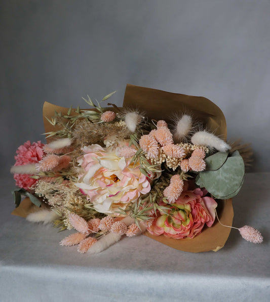 A bouquet of dried flowers wrapped in brown paper, featuring pink, peach, and white blooms, along with dried grasses and green foliage. The bouquet is placed on a  surface against a grey background.
