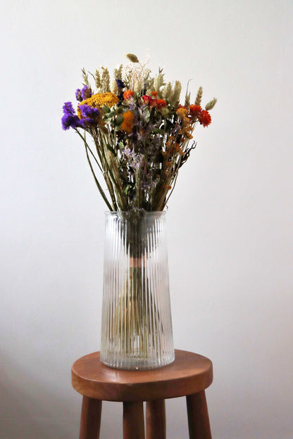 A bouquet of dried flowers in a ribbed glass vase on a wooden stool, featuring purple statice, orange and red strawflowers, and dried grasses against a white background.