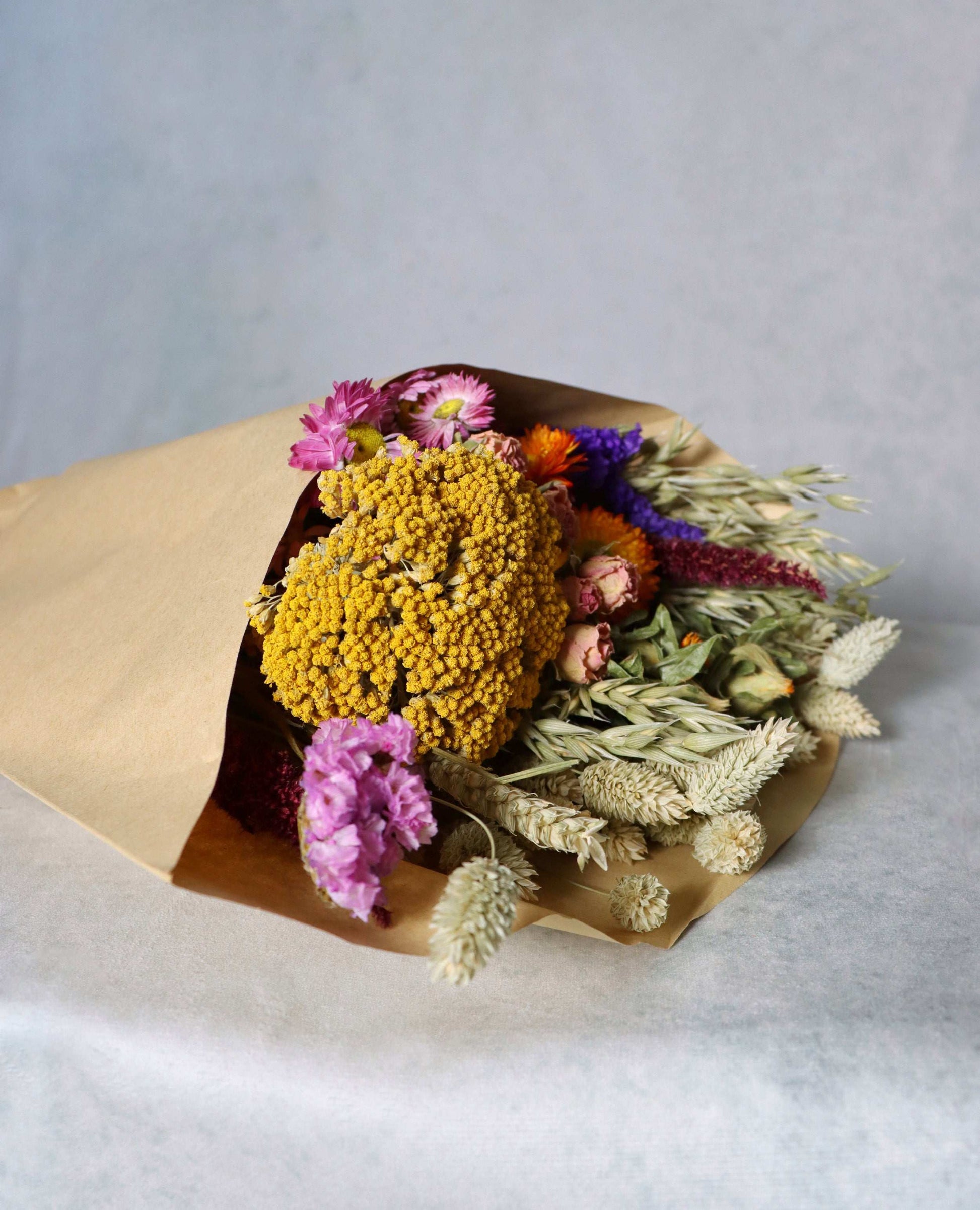 A colourful bouquet of dried flowers wrapped in brown paper, featuring yellow billy buttons, pink strawflowers, orange celosia, and dried grasses on a white surface.