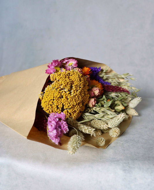 A colourful bouquet of dried flowers wrapped in brown paper, featuring yellow billy buttons, pink strawflowers, orange celosia, and dried grasses on a white surface.