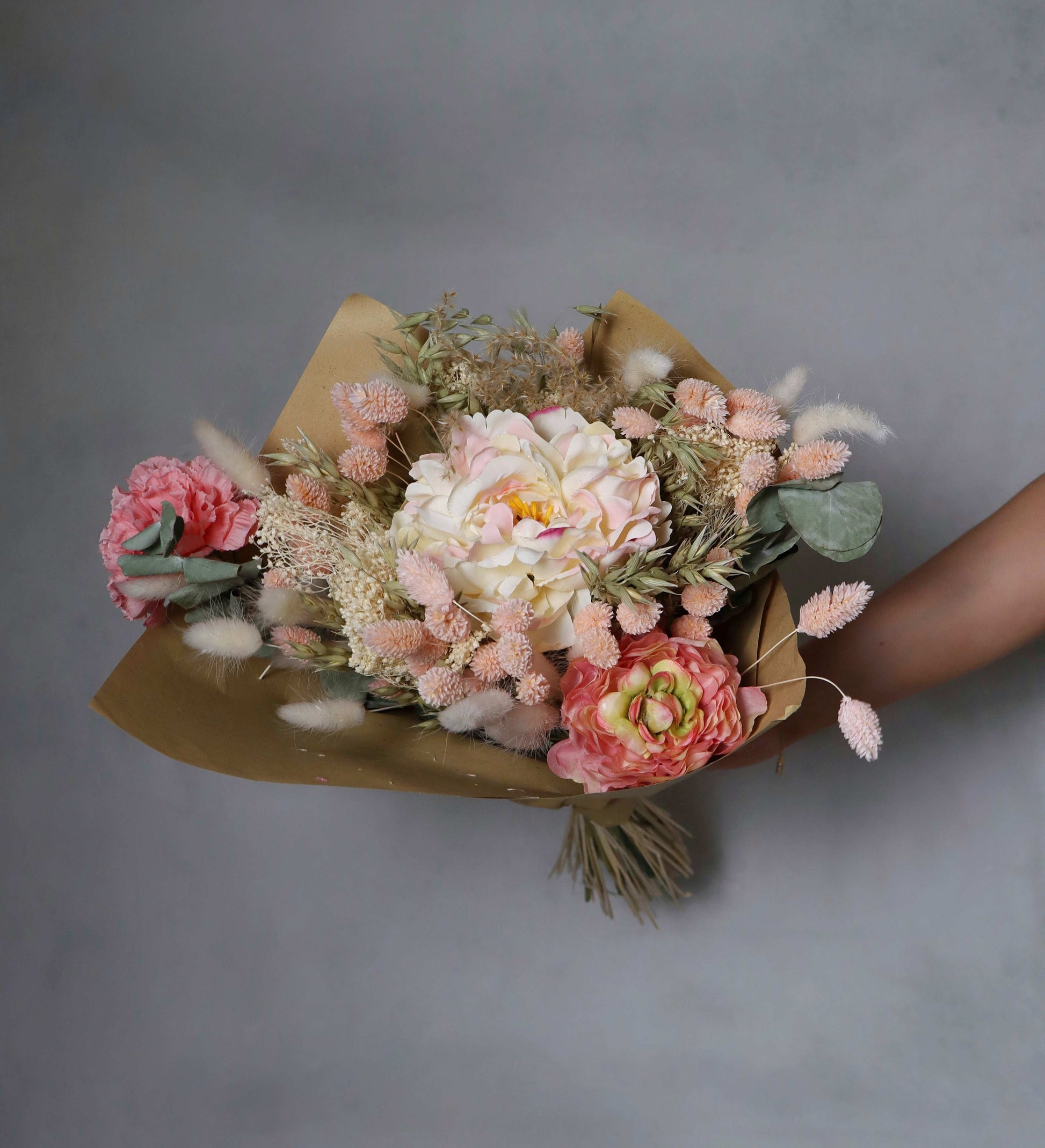A bouquet of dried flowers wrapped in brown paper, featuring pink, peach, and white blooms, along with dried grasses and green foliage. The bouquet is held in a person's hand against a grey background.