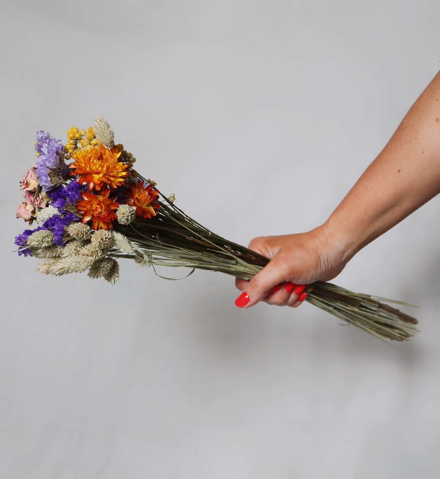 Hand holding a bunch of dried flowers with purple, orange, and beige blooms against a grey background.