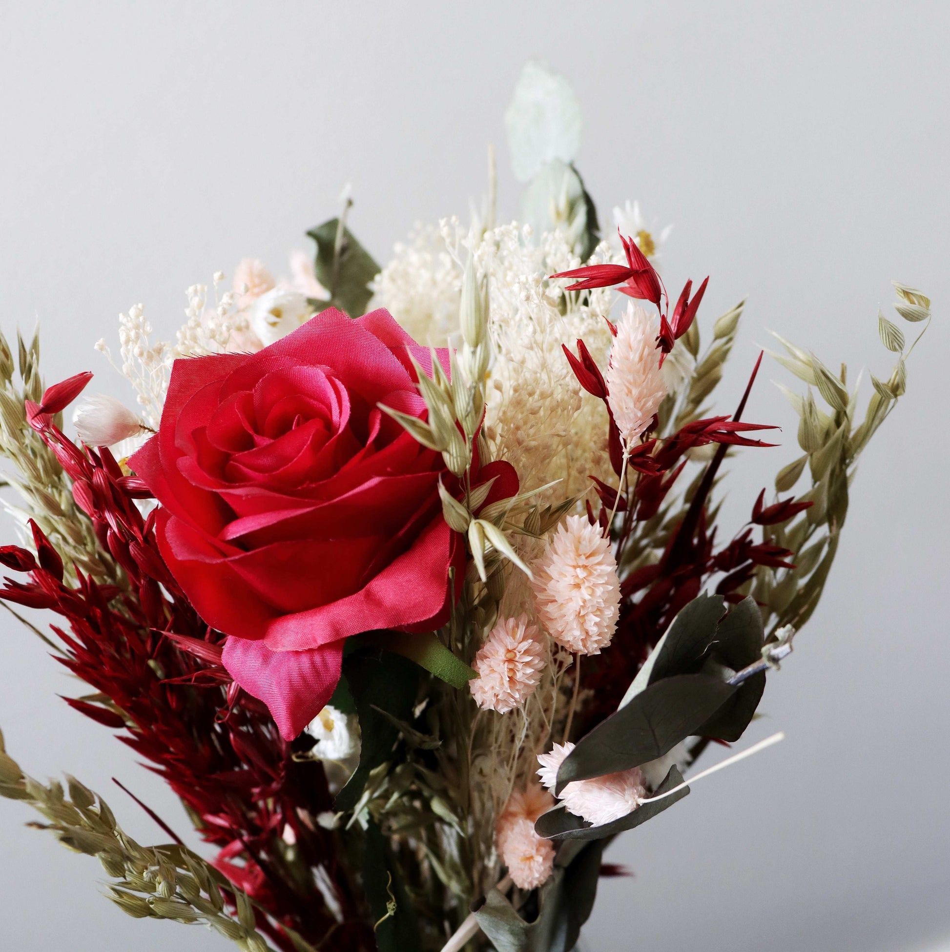 Close up of dried flowers with a red rose, pink blooms, white baby's breath, and green eucalyptus leaves against a grey background.