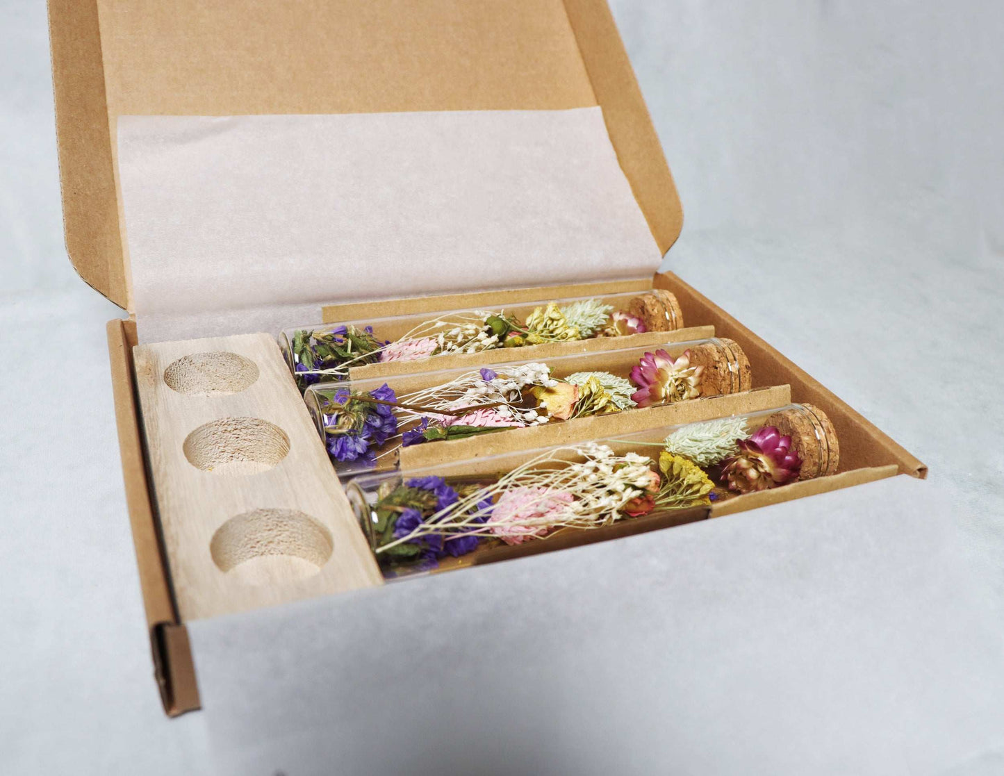 Box containing dried flower bouquet with pink, purple, and yellow blooms, wooden vials, and a stand against a white background.
