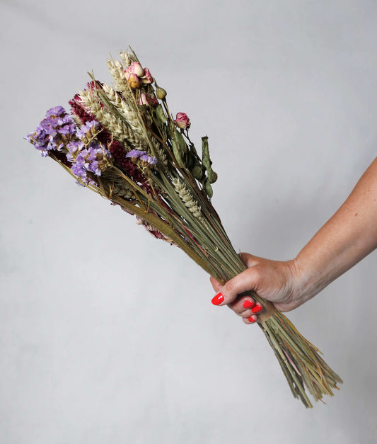 Hand holding a bunch of dried flowers with purple, pink, and beige blooms and wheat stalks against a grey background.